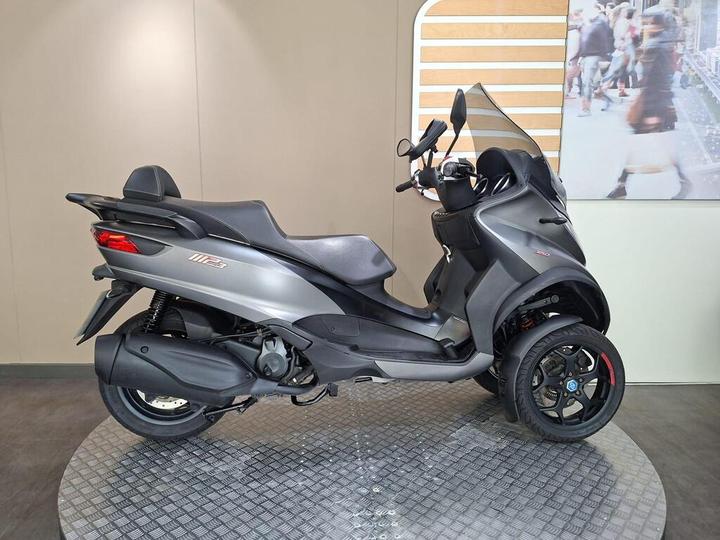 A side view of a gray three-wheeled scooter displayed on a circular platform with a simple light-colored background.