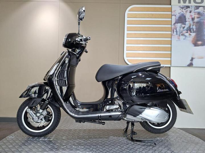 A glossy black Vespa scooter is displayed on a textured metal platform against a neutral background with a faint pattern of people in motion.
