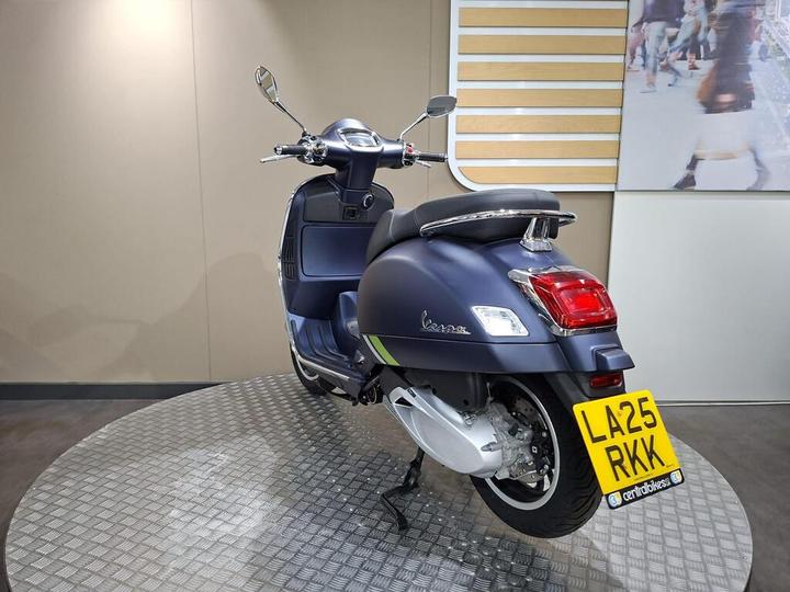 A blue Vespa scooter displayed on a circular, textured platform, with a distinctive license plate L425 RKK visible. The background features a neutral-colored wall.