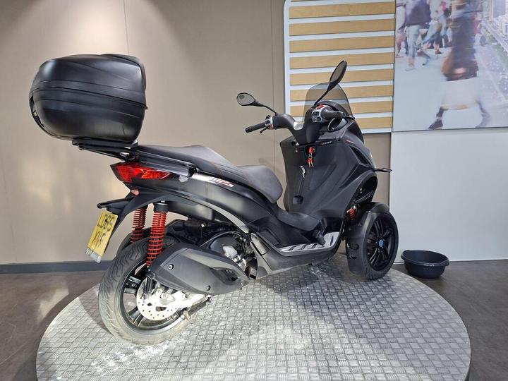 A black three-wheeled scooter with a rear storage box, parked on a circular patterned surface in a modern interior setting.
