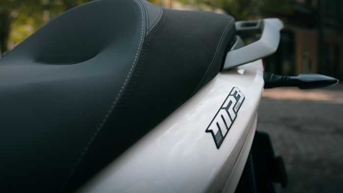 Close-up view of a motorcycle seat featuring a black and gray design, with the model name "MP3" prominently displayed on the white body of the bike. Blurred background of trees and street.