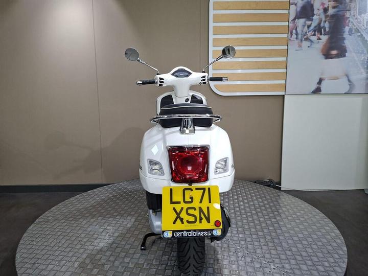 A white scooter viewed from the rear, showcasing the license plate "LG71 XSN," surrounded by a display backdrop and standing on a circular patterned surface.