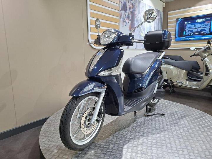 A blue scooter displayed on a circular platform in a showroom, with a digital screen showing information in the background.