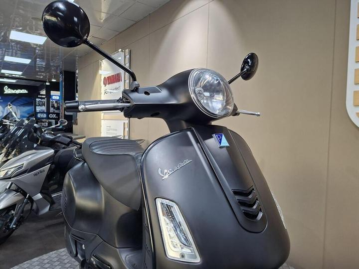 A sleek black Vespa scooter is showcased in a dealership, featuring a prominent front headlight, mirrored handlebars, and a textured seat. The background includes other motorcycles and a display with branding signs.