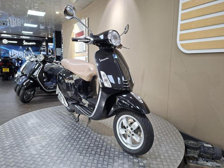 A black Vespa scooter with a tan seat is displayed on a circular platform in a dealership showroom, with additional scooters visible in the background.