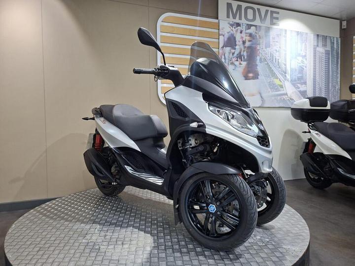 A modern three-wheeled scooter in silver and black, displayed on a circular platform with a textured surface, set against a neutral wall with a "MOVE" sign and a cityscape mural in the background.