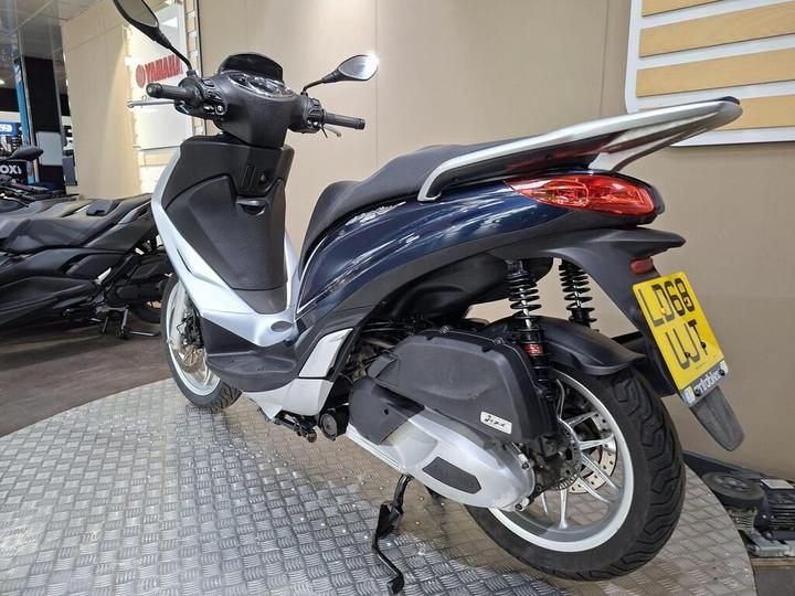 A blue and silver scooter stands on a textured metal surface, showcasing its rear and right side, with the license plate visible. In the background, several dark motorcycles are partially visible, creating a contrasting display.