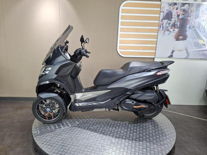 A three-wheeled scooter displayed on a circular platform, featuring a sleek gray design, a windshield, and modern styling accents, set against a neutral background.
