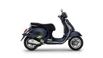 A blue Vespa scooter with green accents is displayed from the side, showcasing its sleek design, chrome wheels, and a comfortable black seat.