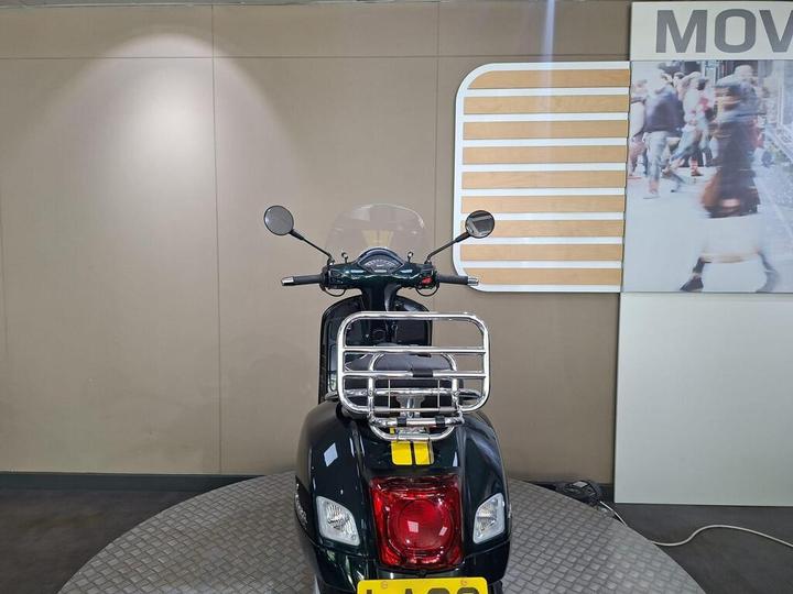 A black scooter is showcased from the rear, featuring a chrome rear rack, a clear windshield, and handlebars with mirrors. The background consists of neutral-colored walls and a large photo of a bustling street scene.