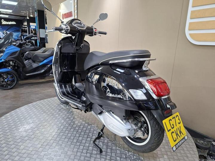 A black Vespa scooter is showcased at a dealership, featuring a sleek design, chrome details, and a visible license plate reading "LG73 CKK." In the background, a blue three-wheeled motorcycle is partially visible. The display area has a checkered floor and modern wall decor.