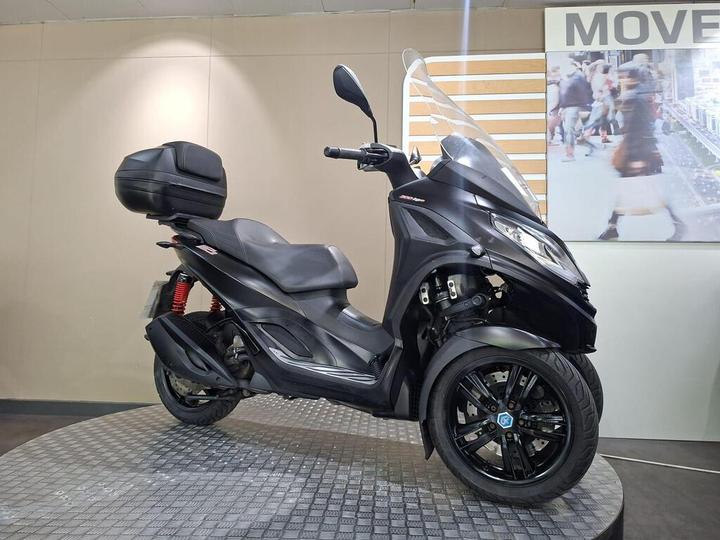 A black three-wheeled scooter with a large windshield and a rear storage box, displayed on a circular platform in a showroom setting.