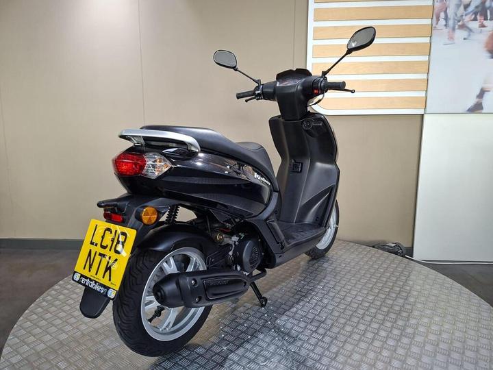 A black scooter displayed on a circular metallic platform, showing its rear and side profile with a visible license plate.