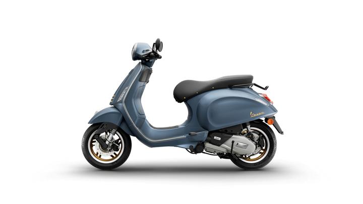 A blue Vespa scooter with a matte finish, featuring a black seat and chrome accents, positioned in profile against a white background.