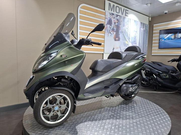 A green three-wheeled scooter displayed on a circular platform in a showroom, with a backdrop featuring a graphic of a city street.
