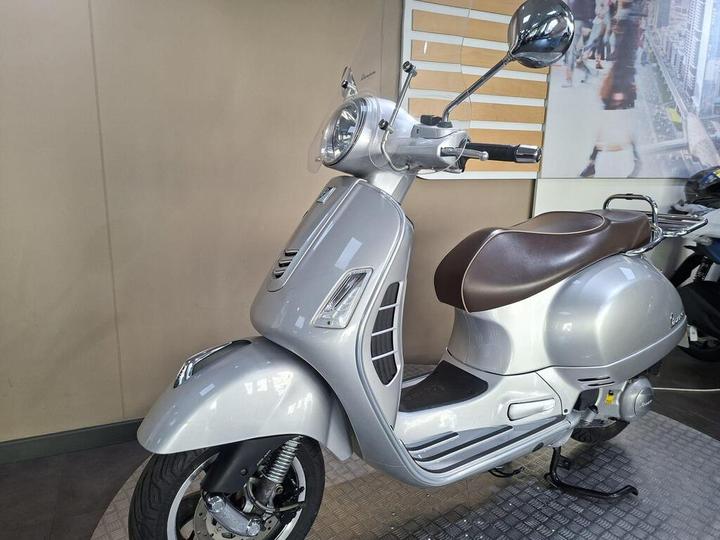 A sleek silver scooter with a brown seat, featuring a round headlight and chrome mirror, parked against a minimalist backdrop.