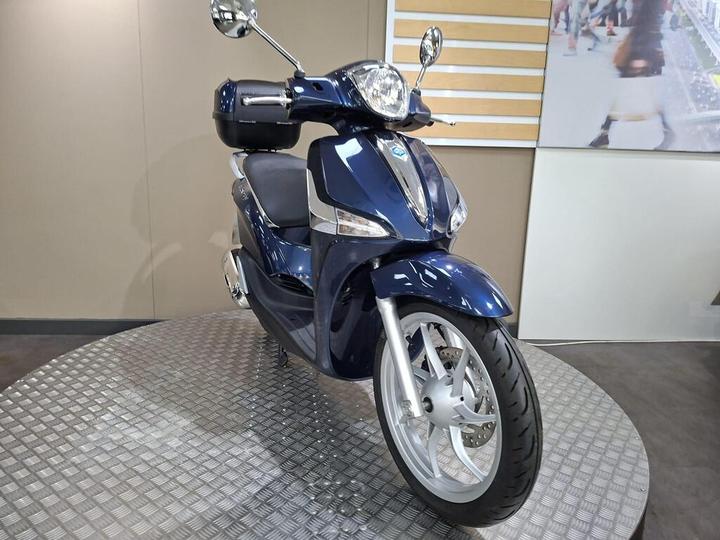 A blue scooter displayed on a round metallic platform with a neutral-colored background.
