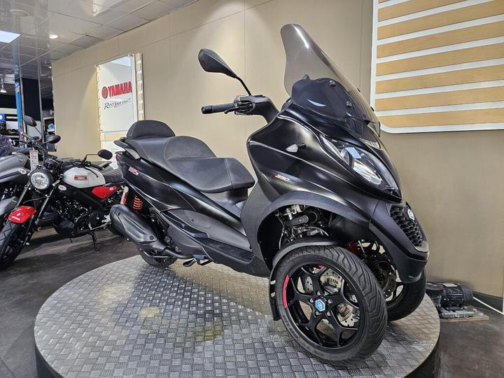 A black three-wheeled scooter displayed on a circular metallic platform in a showroom, surrounded by other motorcycles. The background features a Yamaha sign.