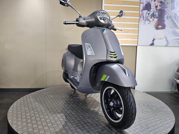 A sleek, gray Vespa scooter is displayed on a circular platform, showcasing its modern design and distinctive features such as circular headlights and green accents. The background is minimalistic with a light-colored wall.