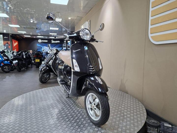 A black Vespa scooter displayed in a showroom, featuring a brown seat, reflecting the interior lights and surrounded by other scooters. The background includes promotional signage and various motorcycles, all on a circular metal platform.