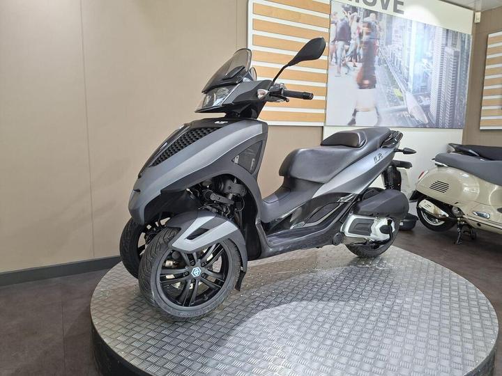 A stylish gray three-wheeled scooter displayed on a circular platform in a modern showroom, with a neutral background featuring a blurred image of urban life and other scooters.