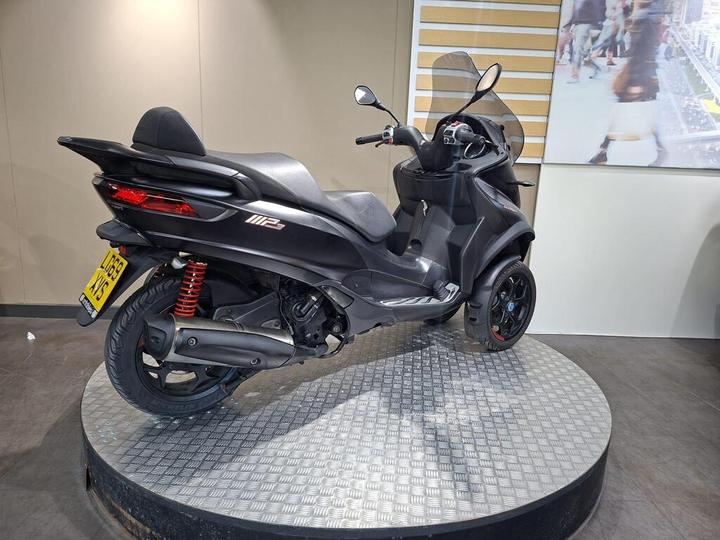 A black scooter with a sleek design is showcased on a rotating circular platform, with visible red suspension springs and a transparent windscreen. The background features a light-colored wall with a large image of people in an urban setting.