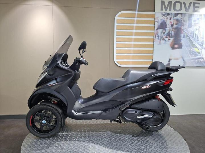 A black three-wheeled scooter on a display platform, featuring a clear windshield, sporty design, and modern wheels, against a light-colored wall with a "MOVE" sign and blurred background of people.