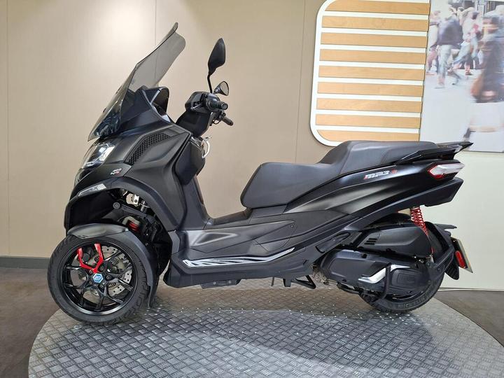 A side view of a black Piaggio MP3 scooter displayed on a patterned gray surface against a neutral background. The scooter features a large windshield, sporty red accents on the wheels, and a sleek design with modern lines.
