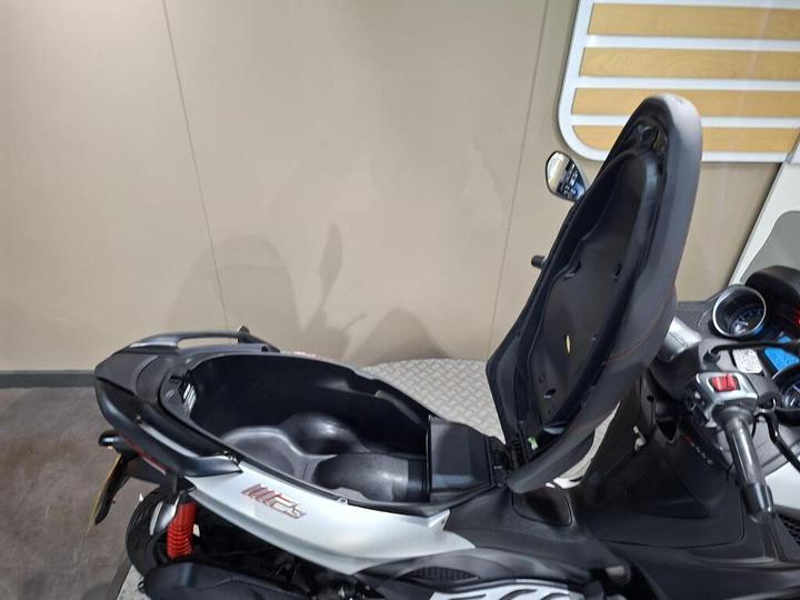 A close-up view of a part of a scooter showing the seat lifted, revealing the storage compartment underneath, with a handlebar visible on the right side. The background is a plain wall.