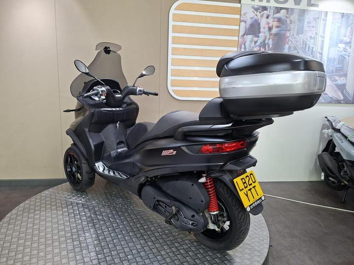 A black and silver scooter with a large storage box on the back is displayed in a showroom. The scooter features a windshield and has red suspension springs, standing on a circular textured platform with a neutral-colored background.