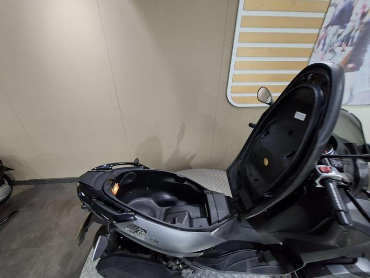 A close-up view of a motorcycle seat with the seat back reclined, revealing a storage compartment underneath. The background features a neutral-colored wall with abstract designs.
