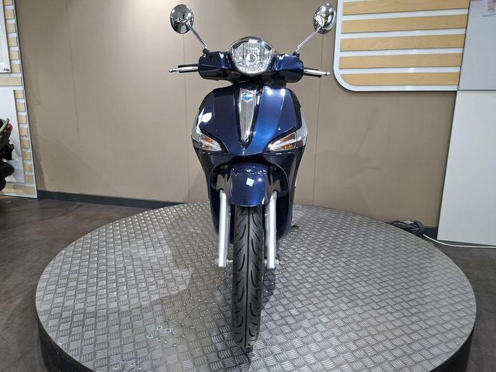 A blue scooter is displayed on a circular platform, viewed from the front, showcasing its sleek design, chrome mirrors, and detailed rim pattern. The background features a neutral-colored wall and visible reflections.