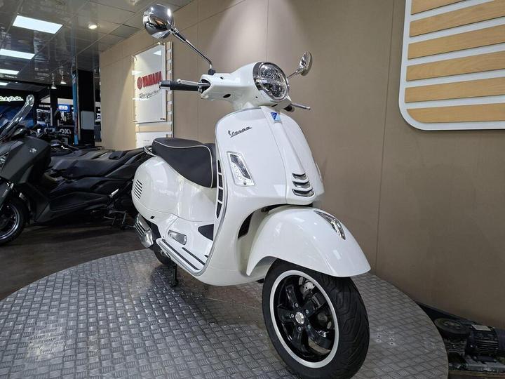 A white Vespa scooter displayed on a circular platform in a showroom, with soft lighting and motorcycles visible in the background.