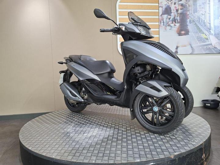 A sleek gray scooter with three wheels displayed on a circular platform in a modern showroom setting. The background features a subtle, light-colored wall and a promotional image of a busy urban scene.
