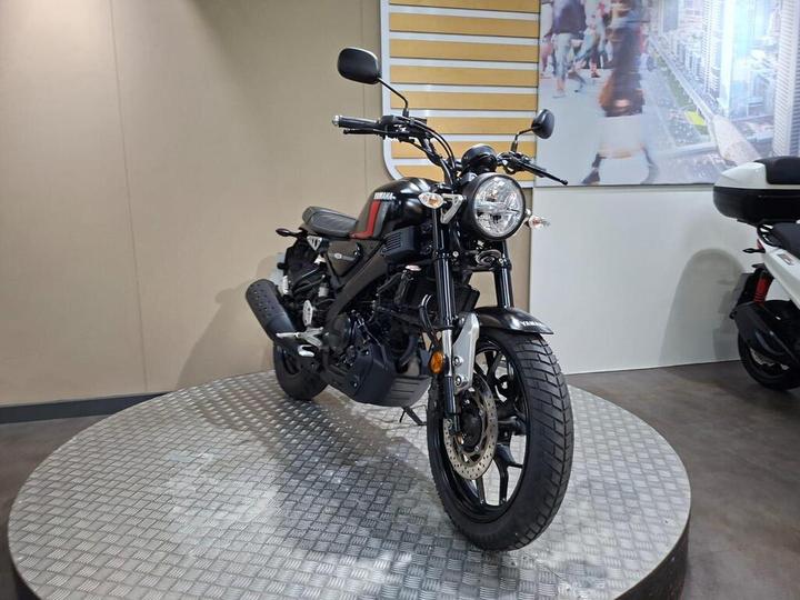 A black motorcycle with a retro design, featuring a round headlight and a minimalist body, displayed on a circular platform in a showroom with a neutral background.