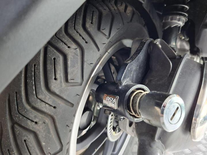 Close-up view of a motorcycle tire with a distinctive tread pattern, showcasing the brake caliper, axle, and a part labeled "Easy Block." The image captures details like dirt on the tire and components of the motorcycle's suspension system.