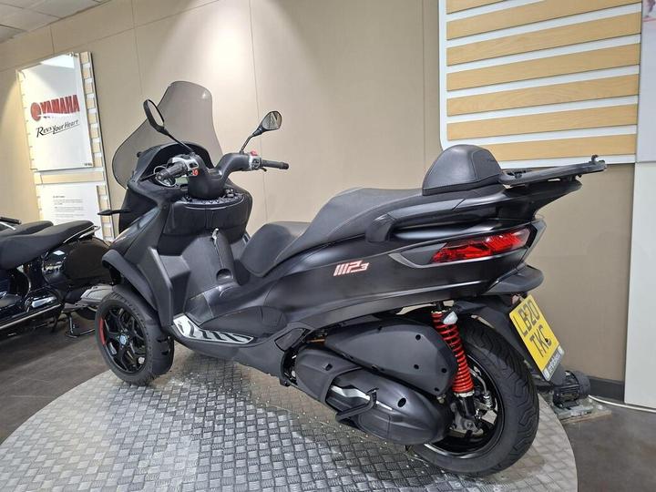 A black Yamaha MP3 scooter displayed in a showroom with a transparent windscreen, showcasing its sleek design and red spring suspension.
