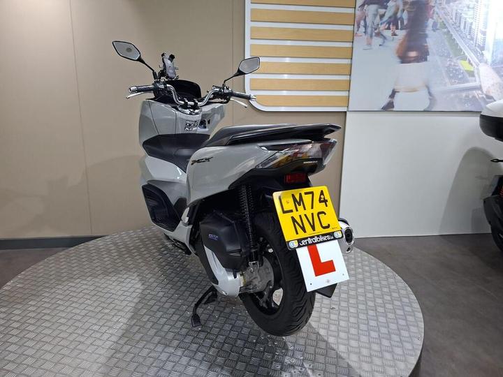 A gray Honda motorcycle displayed on a circular platform, featuring a learner's plate with the registration number LM74 NVC. The background includes a beige wall and a large image of a city scene.