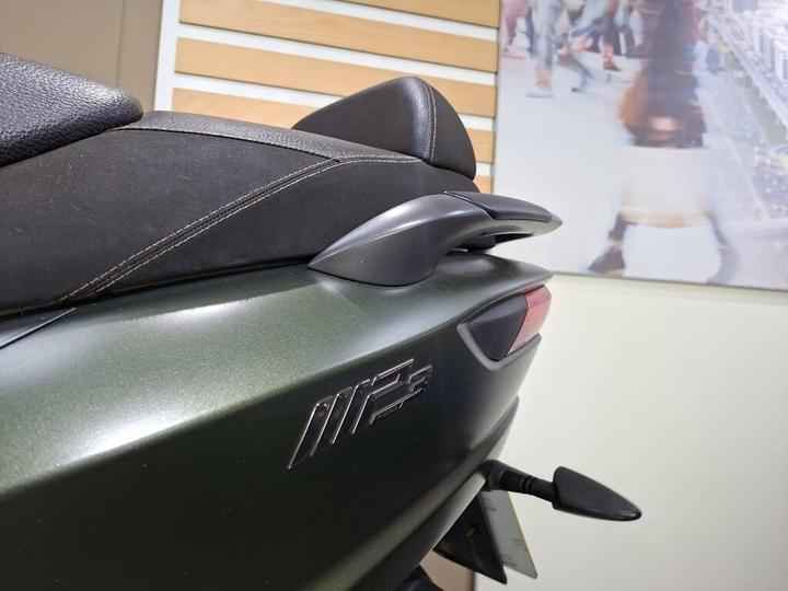 Close-up of the rear section of a motorcycle, featuring a matte black seat and a sleek gray tail piece with the model designation "M F." A blurred background shows a commercial or urban scene.