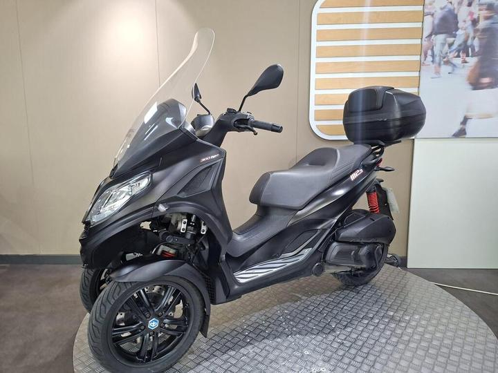 A black three-wheeled scooter displayed in a showroom, featuring a large windshield and rear storage box, with a modern design and sporty wheels.