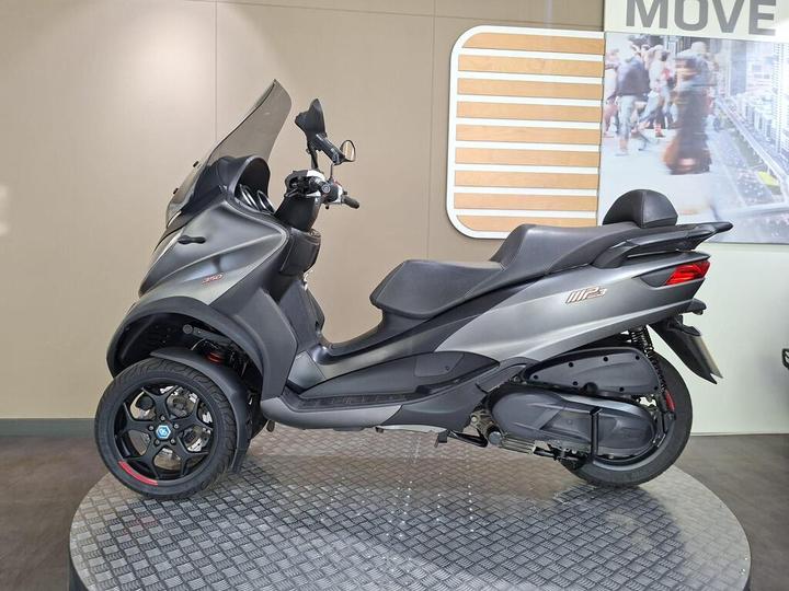 A sleek gray three-wheeled scooter showcased on a circular black platform in a modern display setting. The scooter features a visor, comfortable seating, and sporty wheels, with a background partially visible showing a bustling street scene.