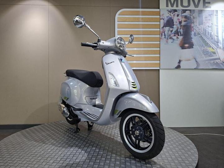 A sleek silver Vespa scooter is displayed on a circular platform, with a minimalist background featuring a cityscape image and the word "MOVE." The scooter showcases modern design elements, including chrome mirrors and a black seat.