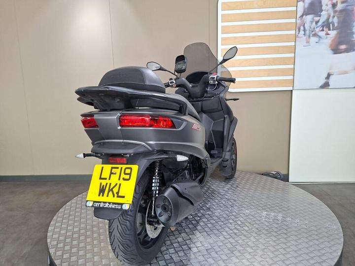 A black three-wheeled scooter with a prominent rear view, featuring a license plate that reads "LF19 WKL." It is displayed on a circular, textured platform with a neutral background.