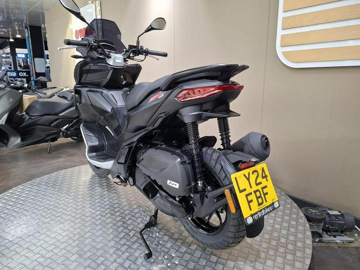 A black scooter on a patterned display, featuring a sleek design, prominent tail lights, and a visible license plate reading "LY24 FBF." In the background, various other scooters and a shop interior are partially visible.