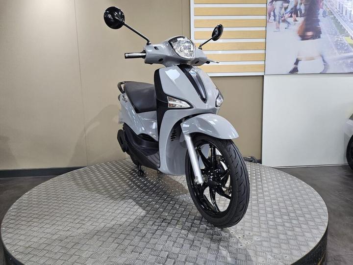 A light gray scooter is positioned on a textured circular platform, showcasing its sleek design and black wheels. The background features a simple, neutral wall.