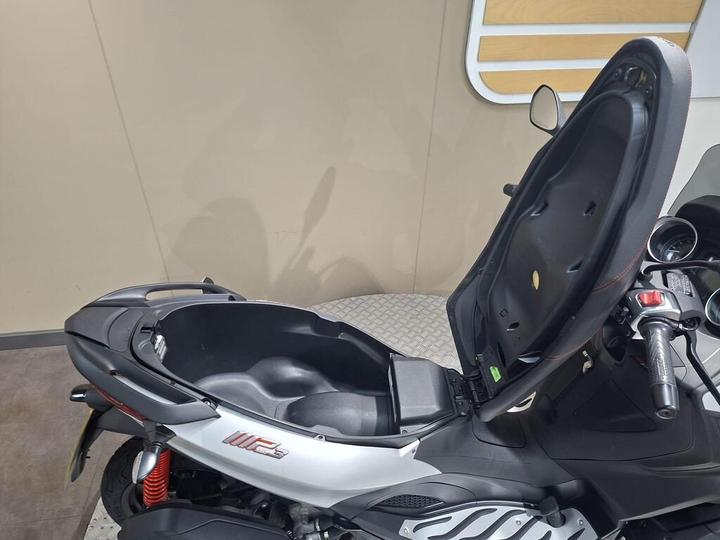 A side view of a scooter with its seat lifted, revealing a storage compartment beneath. The scooter features a sleek design with a silver and black color scheme, and is positioned against a neutral background.