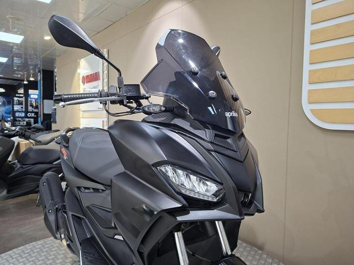 A close-up view of a sleek black Aprilia scooter, featuring modern headlights, a tinted windscreen, and handlebars with control elements, displayed in a well-lit showroom with other motorcycles in the background.
