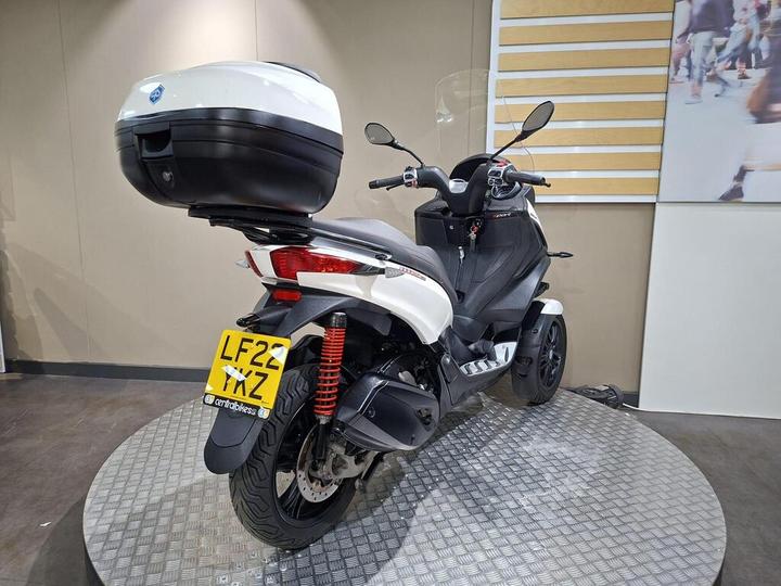 A rear view of a white and black three-wheeled scooter with a top case, displayed on a circular platform. The scooter shows a distinctive design with visible suspension and license plate reading "LF22 YXZ."