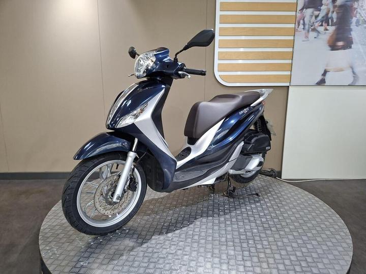 A modern blue and silver scooter displayed on a circular platform, with a minimalist background featuring a grid pattern and light-colored walls.