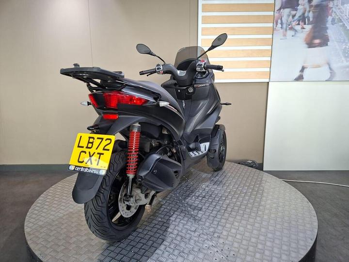 A black scooter displayed on a circular platform, showing the rear view with a visible license plate reading "LB72 CXT" and red suspension springs. The background features a neutral wall and a partially visible image of people walking.